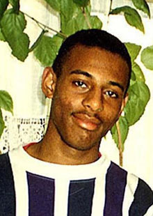 Photograph of a young Afro-Caribbean male, cropped to show his chest and head. He has black hair, shaved very short, and a slight moustache. He is wearing a navy and white vertically-striped crew neck shirt. He is standing in front of a large indoor plant.