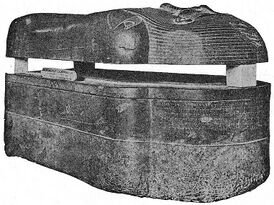 A dark stone coffin viewed laterally, the coffin lies on the ground, the trough and lid are separated with wedges.
