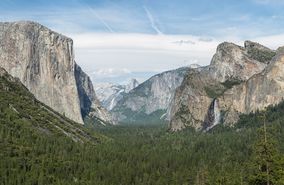 Mountain valley with sheer cliff on its left side, and a waterfall cascading into its right, with a clear blue sky above and many green trees below