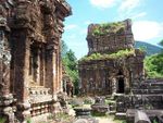 Ruins of buildings of red stone with niches and sculptures. The roof of one of the structures is partially covered in grass.