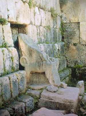 A moss covered stone throne with sphinxes and cloven feet. Behind the throne are moss-covered bare ashlar walls.