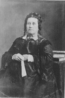 Portrait of an unsmiling, middle-aged woman in a voluminous dress. Her hair is piled on the back of her head, with ringlets near her ears. She holds a fan in her hands.