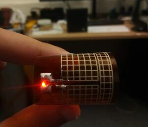 A printed rectenna lighting an LED from a Powercast 915 MHz transmitter, flexible meshed antenna bent with a red LED light