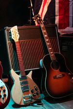 Two guitars lean against an amplifier. The left guitar is electric, and cream-coloured. The right guitar is acoustic, and brown and black in colour. A flag of Great Britain and a second amplifier are visible in the background, and part of a red electric guitar is visible to the left.