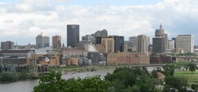 Nearer to the camera, a park area with trees and grass. Behind that is a river which crosses the image. Across the river is a medium-sized downtown with mid- and high-rise building primarily gray and beige in color. The sky is clear with some cloud cover.