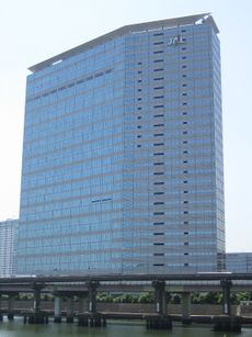 A modern multi-storey building in blue and grey colour, with Japan Airlines' "JAL" logo on the top right, there are blue sky on the background and a highway bridge in the foreground
