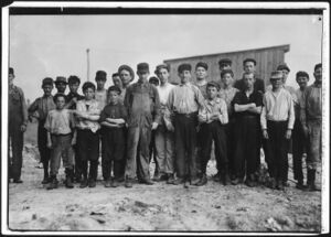 20–25 males facing camera in a row, mostly children. Ages vary. They are in work clothes. Some are dirty. Some wear caps. They stand on dirt with a wooden building behind them.