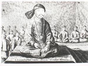 Black-and-white print of a man with small eyes and a thin mustache wearing a robe, a fur hat, and a necklace made with round beads, sitting cross-legged on a three-level platform covered with a rug. Behind him and much smaller are eight men (four on each side) sitting in the same position wearing robes and round caps, as well as four standing men with similar garb (on the left).