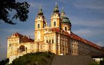 A large oranate church with an enclosed forecourt. The west façade is topped by two towers and the transept crossing is topped by a large round tower. The entire building is decorated with horizontal white and yellow bands.