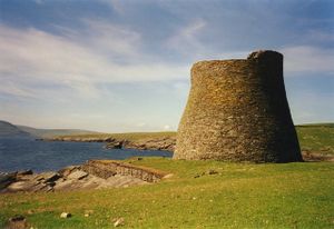 Green fields lie to the right of a body of dark blue water. At the water's edge there is a tall, round, stone tower, possibly unroofed.