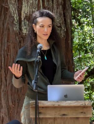 Jay Graber is giving a talk while outside, holding a phone behind a podium. There is a microphone on a stand and a laptop in front of her.