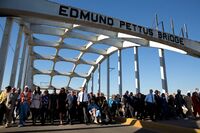 Large group of people walking together on bridge roadway under bridge arch saying "Edmund Pettus Bridge"