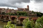 A distant view of a small city in the background and a narrow bridge hidden by a few trees along a river.