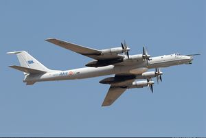 Pale grey aircraft with huge four propellers and swept back wing in flight towards right of camera.