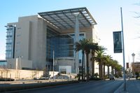 Exterior view of a beige building composed largely of windows. A large overhang attached to the building is supported by a single pillar.