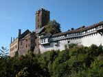 A castle perched along the edge of a wooded hill. The castle has grown in several stages and consists of sections in dark stone, lighter stone, white plaster and half-timber.