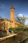 A three story pale red brick building along the left side of a small river. The building has a bell tower rising out of the middle of it.