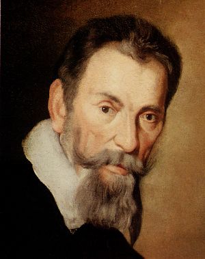 Head of a heavily bearded short-haired man with a serious expression, leaning slightly forward and facing semi-right, although his eyes look straight ahead. A white collar over a dark coat or cloak is also visible.