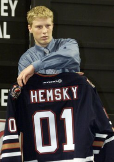 Young man with clean-cut reddish-blonde hair and a blue business shirt and tie, holding up a dark athletic uniform reading "HEMSKY 01" in large white letters