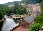 Several red brick factory buildings on the banks of a river which makes a hairpin turn. The buildings are surrounded by mountains and forest.