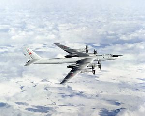 Top perpendicular starboard view of four-engine propeller-driven aircraft in-flight. The pale gray/white aircraft تحلق فوق أرض شبه بيضاء.