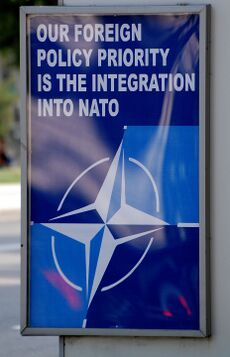 A blue poster at a bus stop with the NATO logo and the words "OUR FOREIGN POLICY PRIORITY IS THE INTEGRATION INTO NATO" in white.